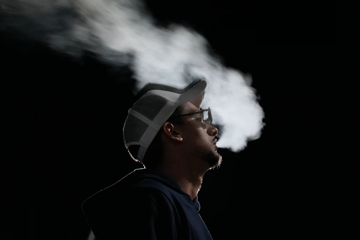 Retrato de perfil en clave baja: chico con gorra blanca y gafas exhalando una nube de humo iluminada a contraluz